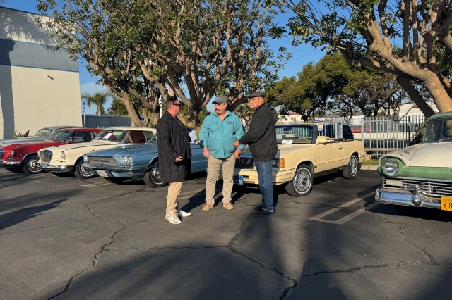 Three men standing in a parking lot in front of their classic cars, 